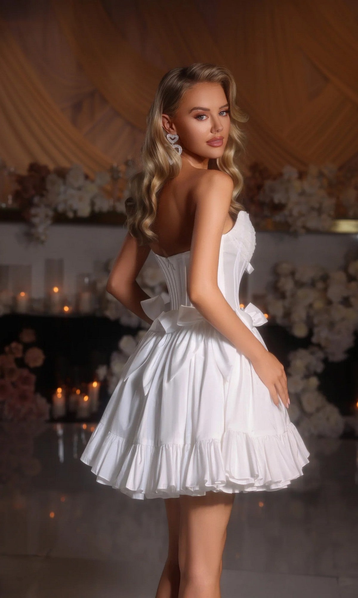 A woman wears the Portia and Scarlett PS25700 short homecoming dress, featuring a white strapless fit-and-flare design with a ruffled skirt. She stands indoors near candles and white flowers, facing slightly away.